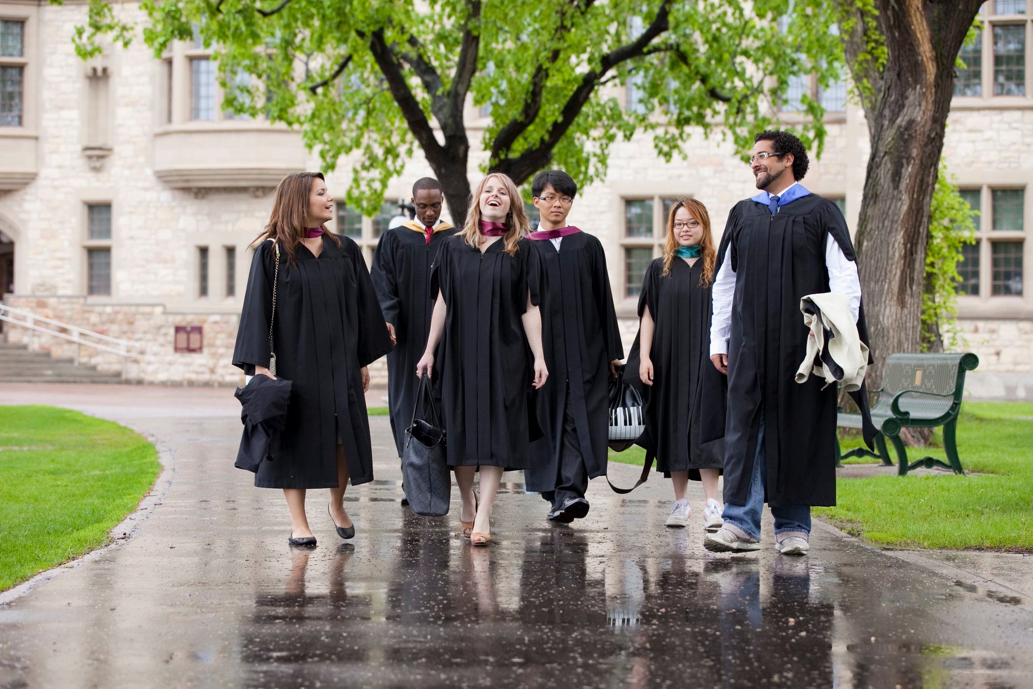 A photo of six young people of different ethnicities, 3 males and 3 females, in graduation gowns walking on a university campus.